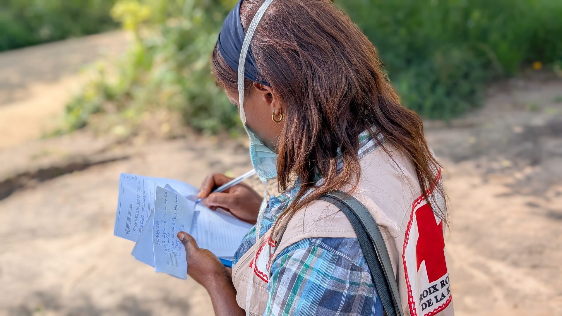 Photograph of a person wearing a mask and a Red Cross vest, writing handwritten notes on a piece of paper. The context suggests medical or humanitarian work outdoors, possibly documentation or data collection.